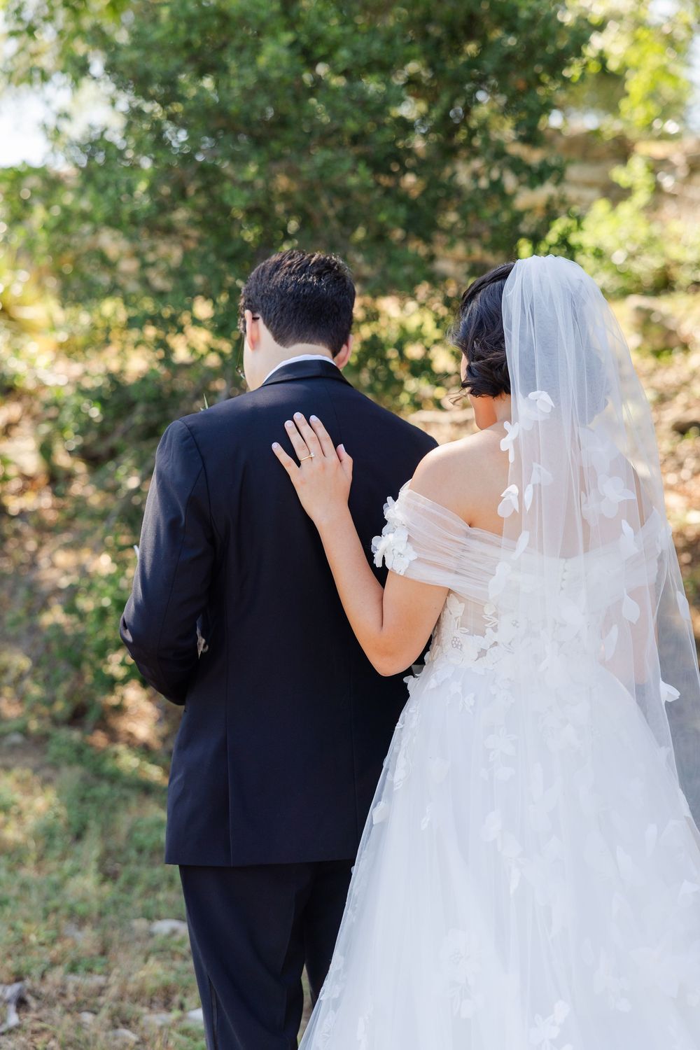 A bride and groom photographed from behind with the bride's hand resting on the groom's back.