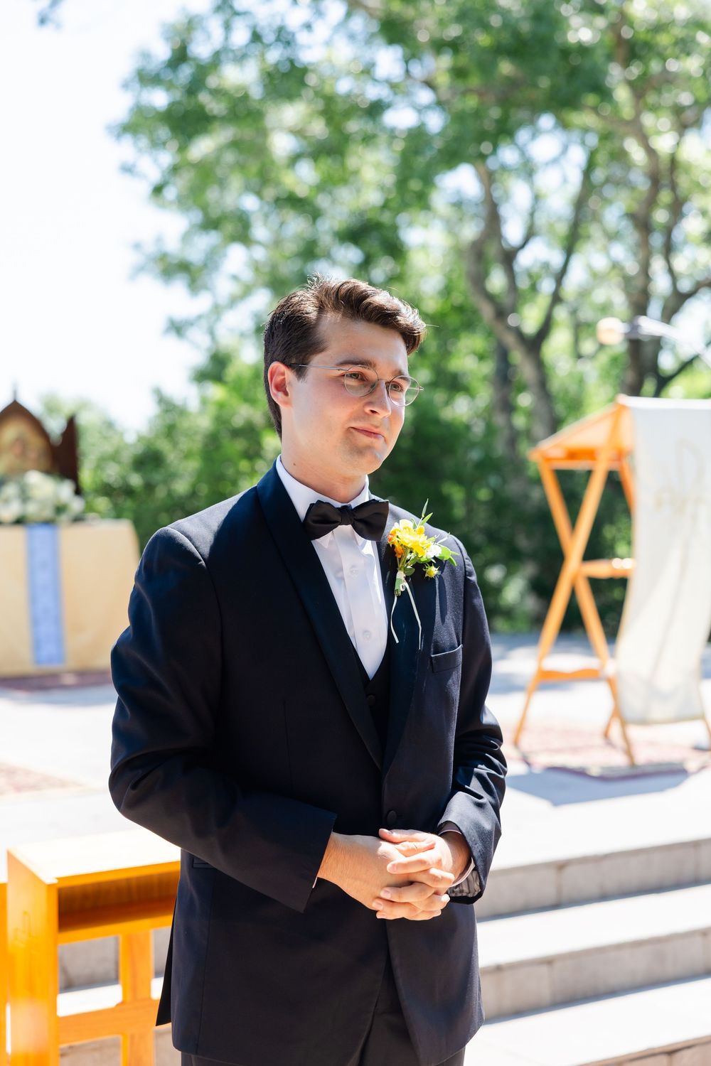 A groom in a classic black tuxedo with a yellow boutonniere stands at an outdoor wedding venue.