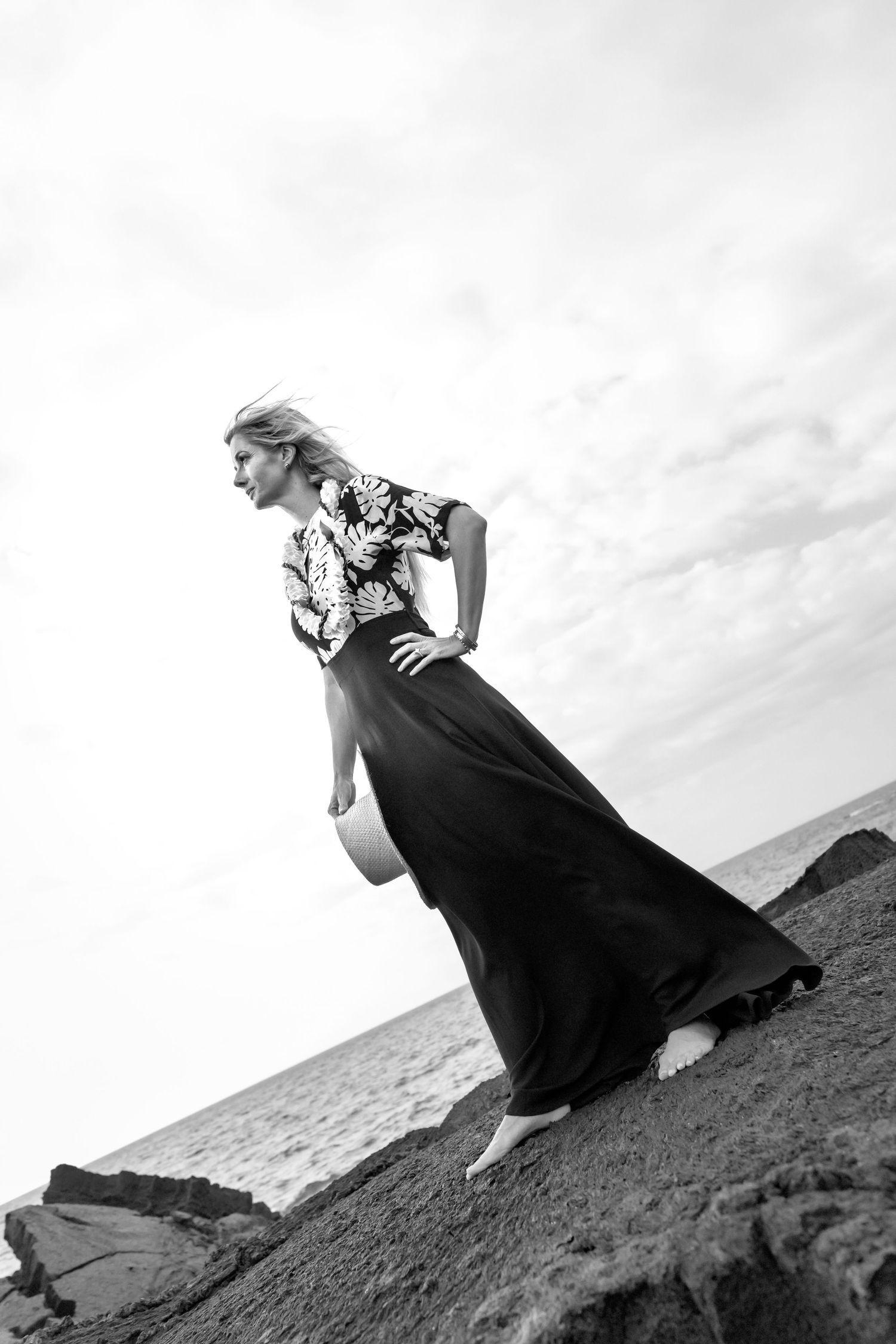 A dramatic black and white portrait of a person in a flowing dark dress posing on rocky coastal terrain against a cloudy sky.
