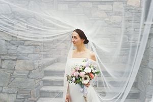 Person in a white off-shoulder wedding dress holds a pink and white floral bouquet against a stone wall backdrop.
