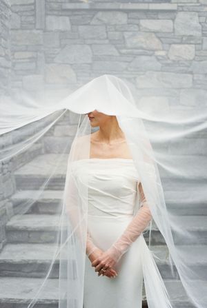 Ethereal bridal portrait showing a classic white wedding gown with sheer veil against a stone wall backdrop.