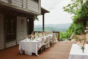 An elegant outdoor dining setup with white linens and chairs on a wooden deck overlooking mountain views.