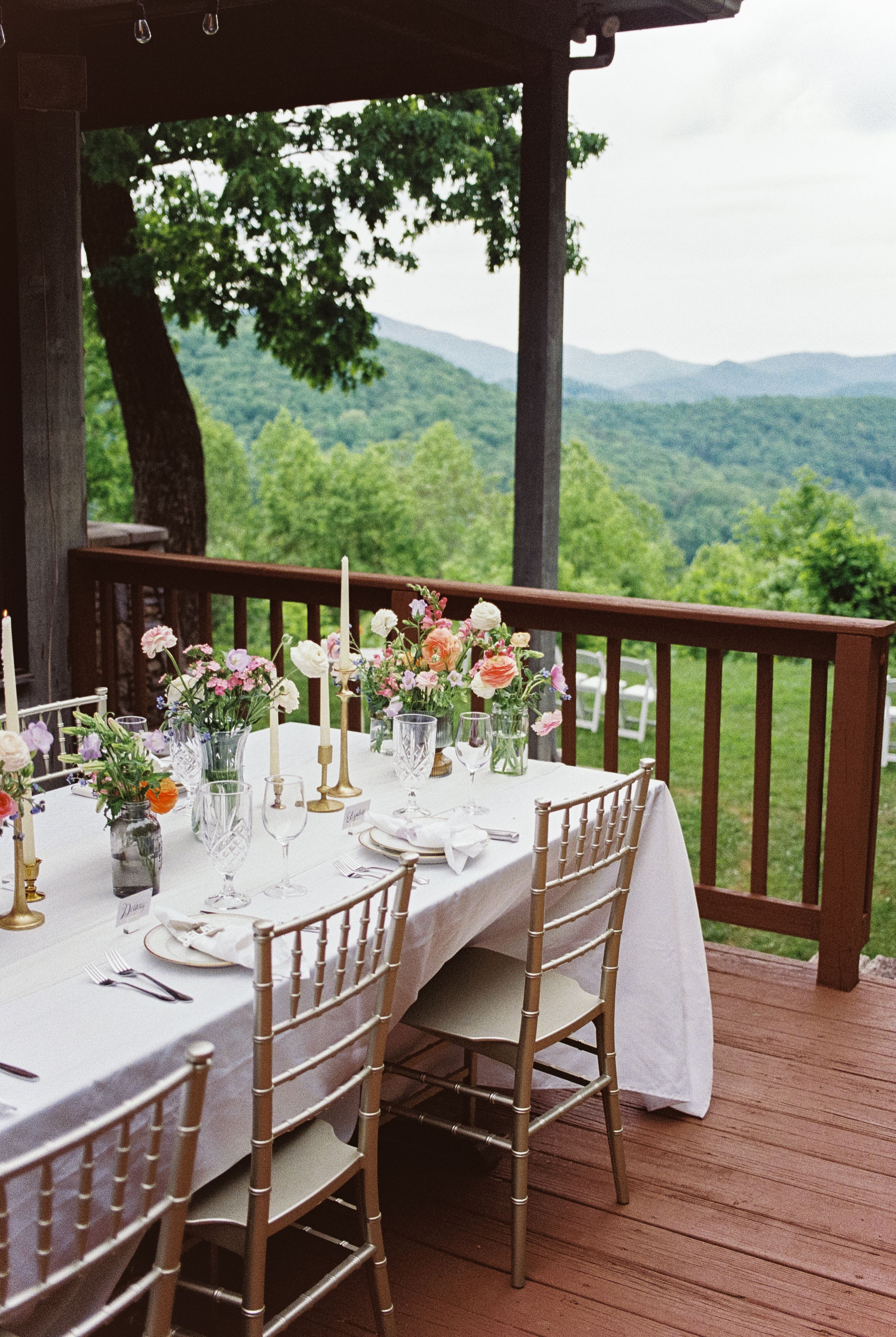 Elegant table setting on a mountain view deck with floral centerpieces, white linens and gold chiavari chairs.