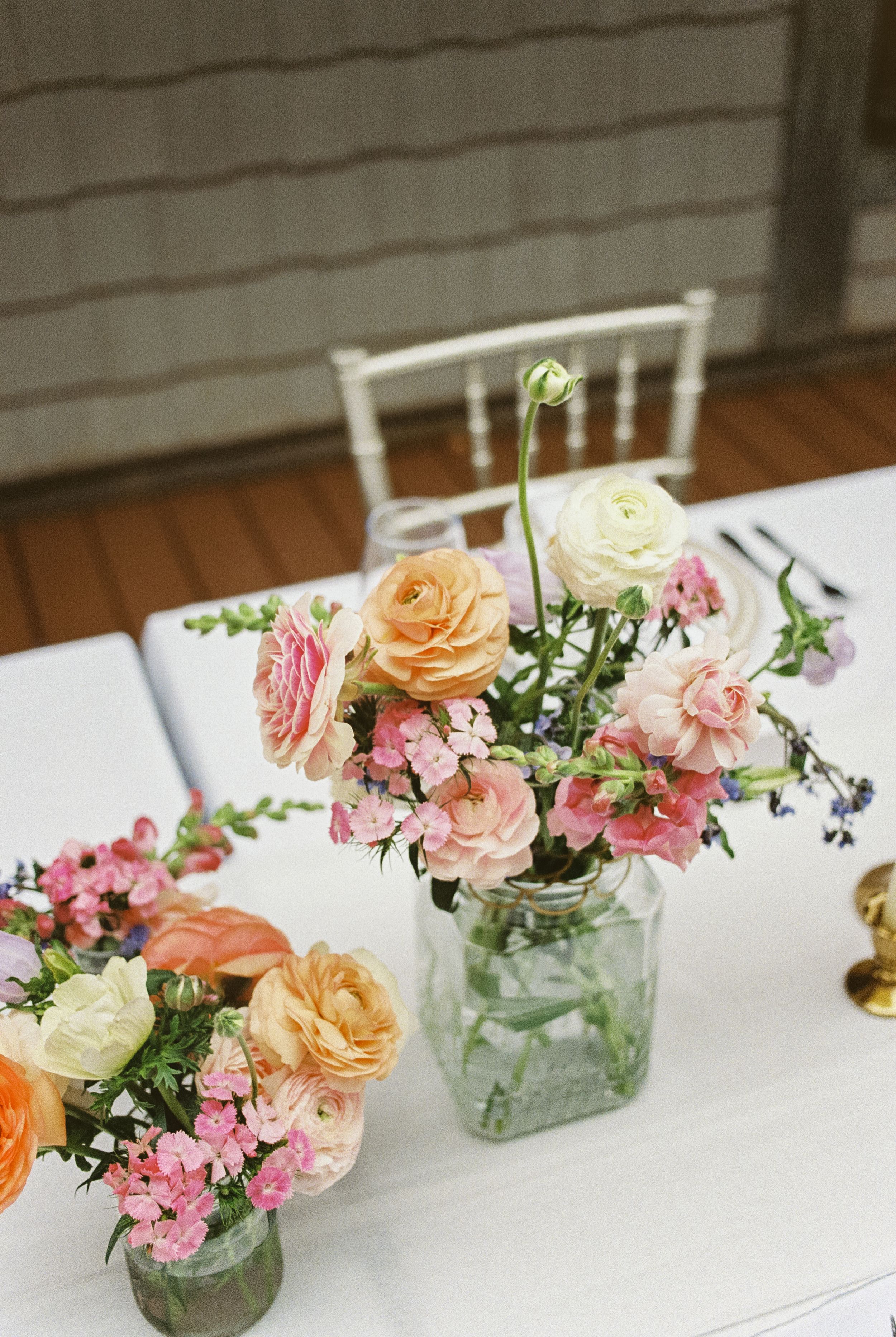 Mason jar centerpieces filled with fresh peach, pink and cream garden roses and wildflowers on white tablecloth.