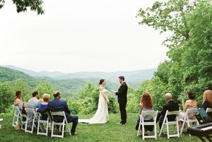 A mountaintop wedding ceremony setup in a lush green setting with white chairs and scenic mountain views in the background.