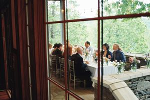 Guests sit at dining tables near large windows overlooking lush greenery at an elegant event venue.