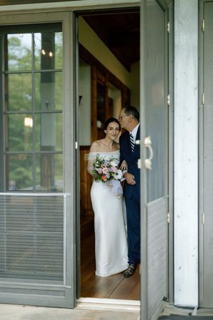 A person in a white wedding dress holds a floral bouquet while standing in an open doorway with gray-blue screens.