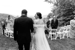 A black and white photo of a bride and groom walking together at an outdoor wedding ceremony with white chairs.