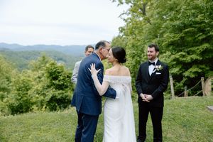 An outdoor wedding ceremony takes place on a grassy hilltop with rolling mountains and green trees in the background.