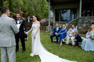 An outdoor wedding ceremony takes place on a grassy lawn with guests seated and watching the exchange of vows.