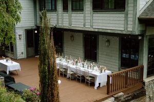 Long white dining table set up for an outdoor reception on a wooden deck next to a rustic style building.