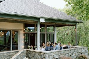 Guests gather on a covered stone patio of an elegant event venue with glass windows and green trim surrounded by trees.