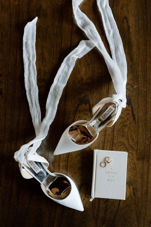 White bridal shoes with ribbons displayed next to a small wedding book on a dark wooden surface.