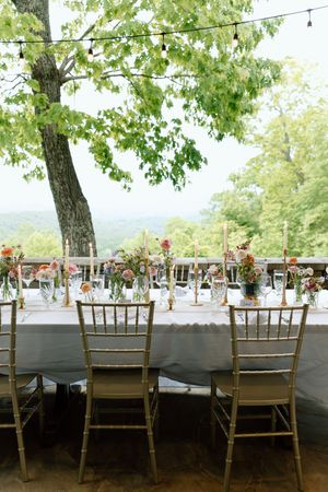 Elegant outdoor reception setup with gold chiavari chairs and floral centerpieces under string lights and tree canopy.
