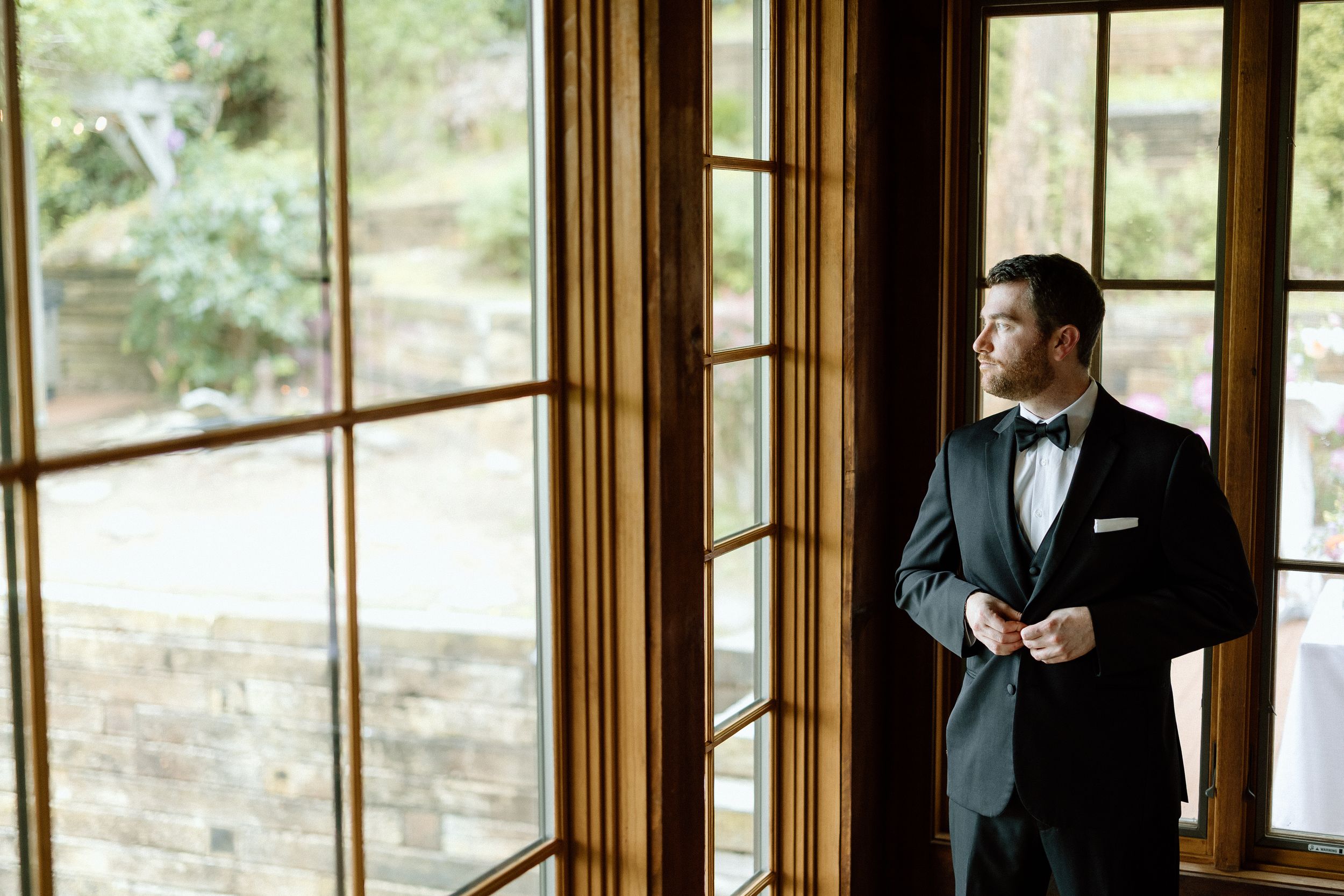 Someone in a black tuxedo and bowtie stands contemplatively by large window panes in a wooden-framed room.
