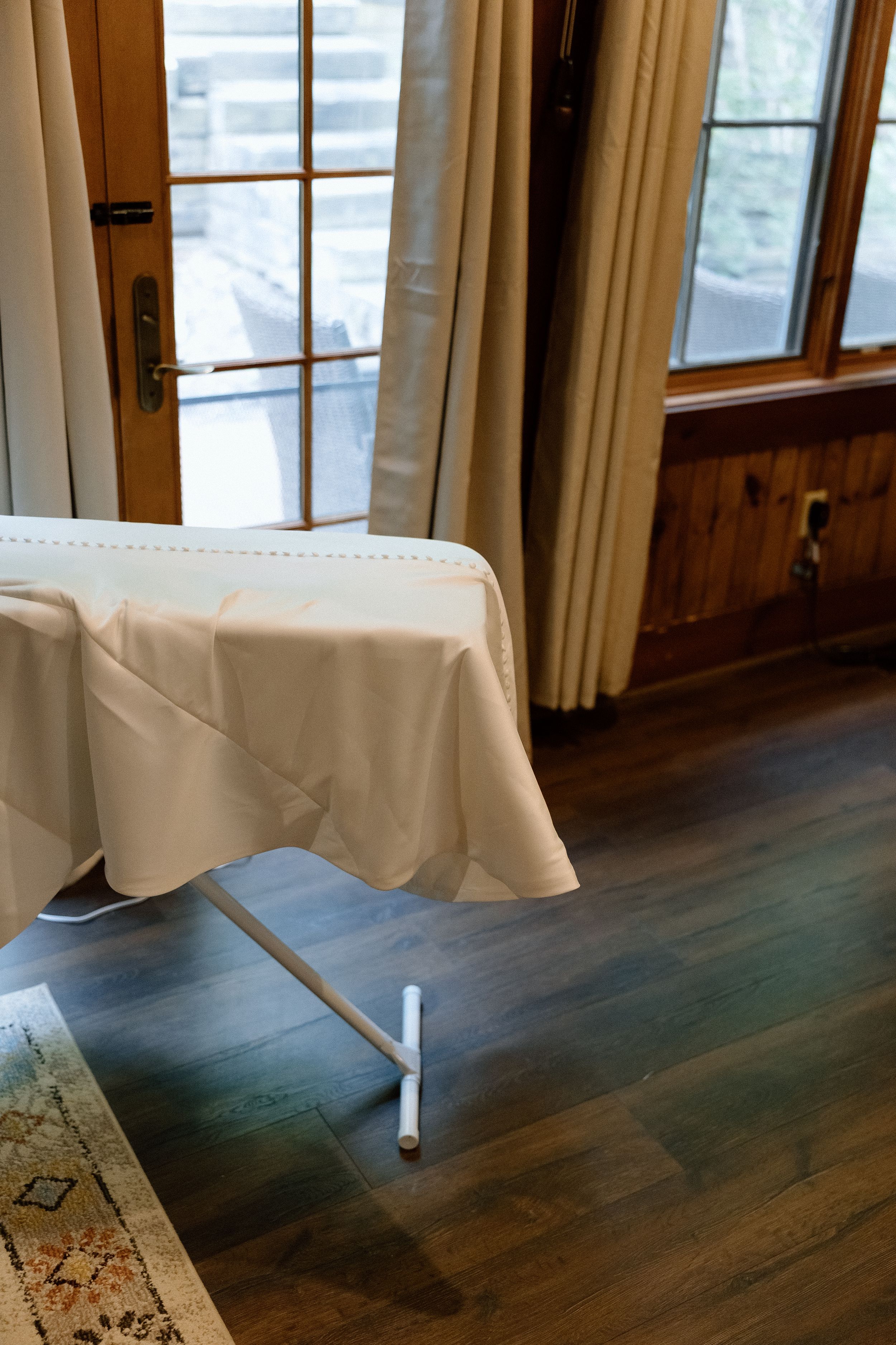 A white tablecloth covers a folding table on dark hardwood flooring near windows with beige curtains.