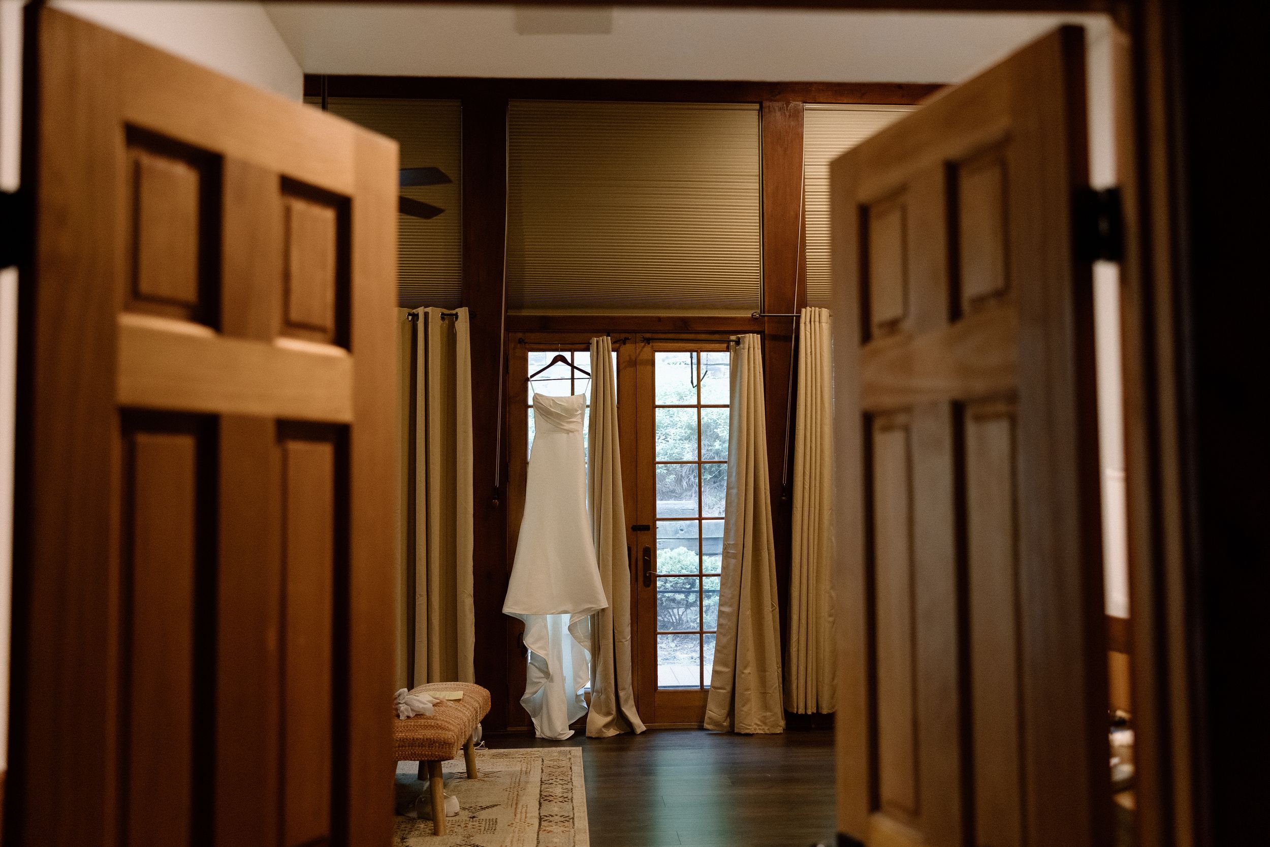 Through open wooden doors, a white wedding dress hangs ethereally between windows in a sunlit room.