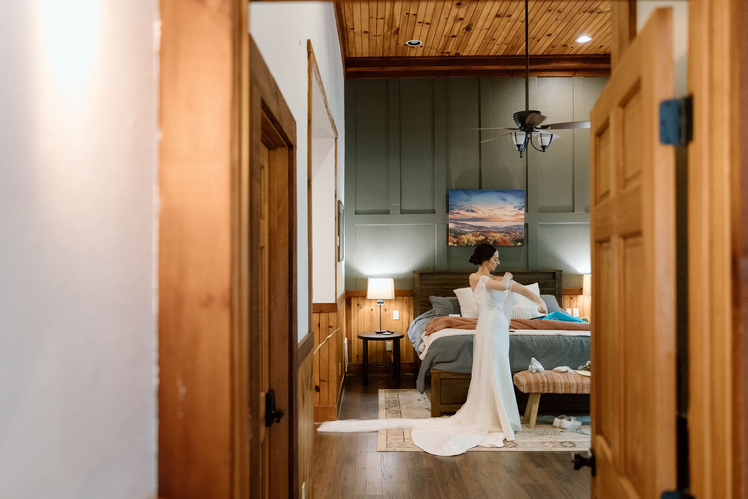 A flowing white wedding dress hangs in the doorway of a cozy cabin bedroom with rustic wood details and soft lighting.