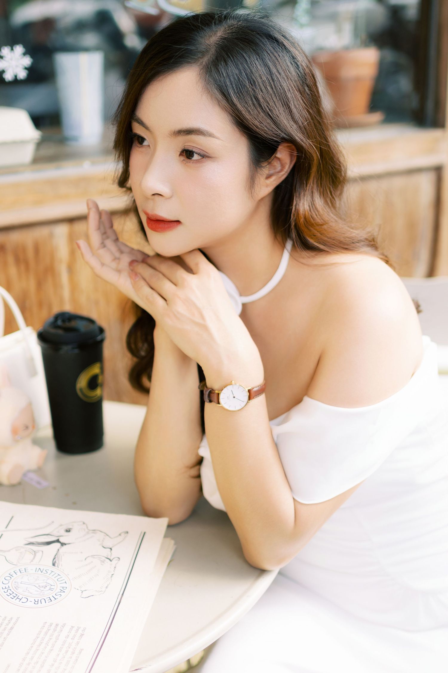 A woman in an elegant white off-shoulder dress poses gracefully outside a cafe with rustic wooden architecture.
