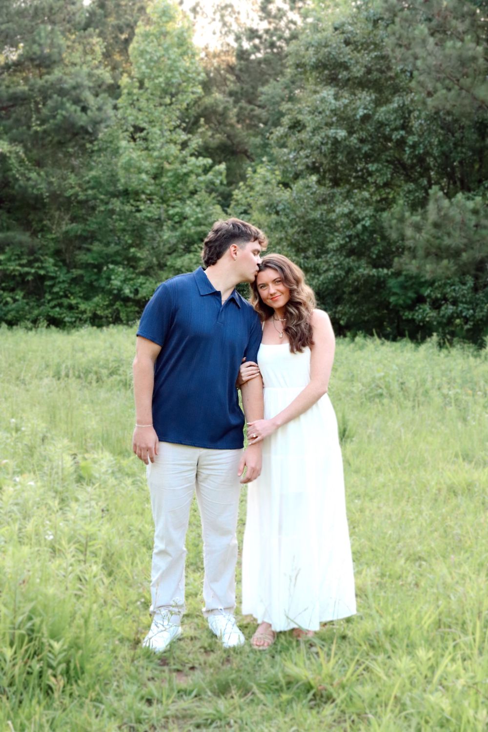 A couple stands together in a grassy meadow with green trees in the background.