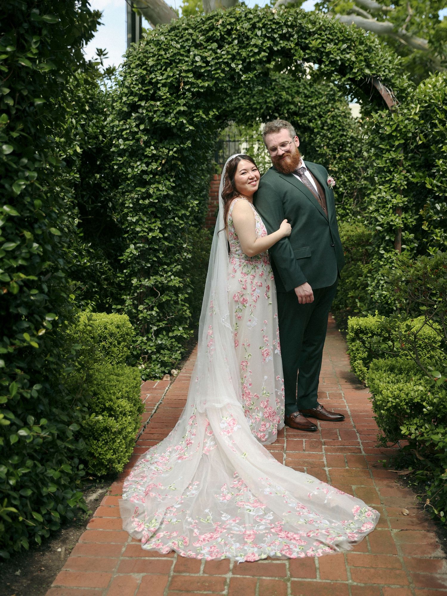 A couple poses on a brick path under a green garden arch, with the bride wearing a floral embellished wedding gown.