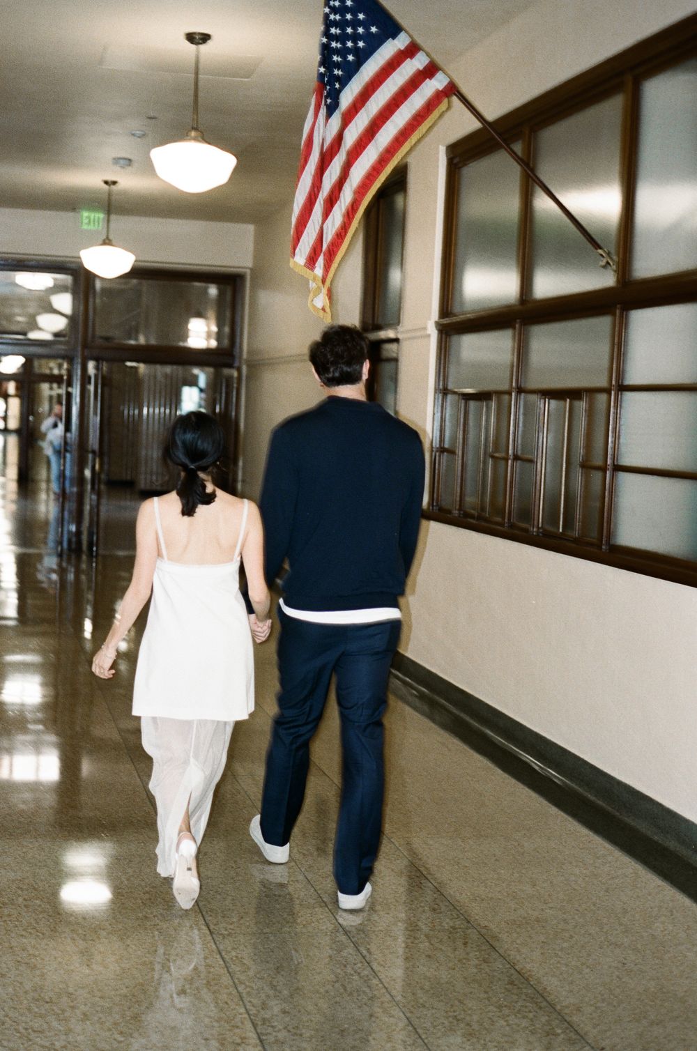 Two people walk down a long institutional hallway with American flags mounted on the walls.