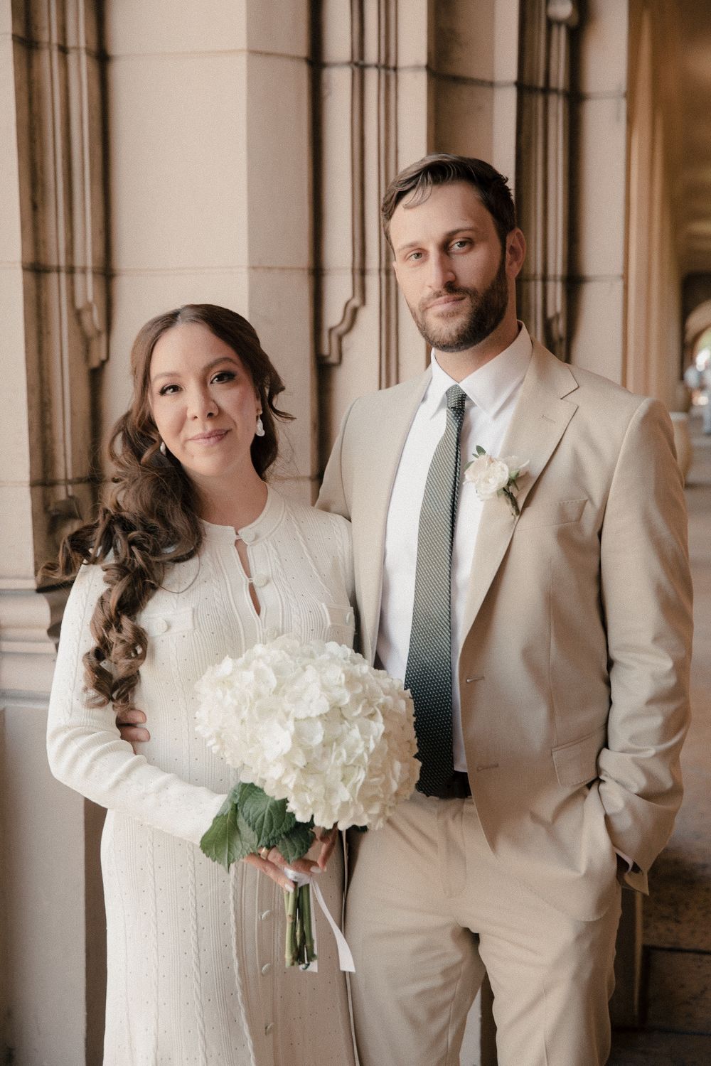 Wedding couple poses in elegant archway wearing beige suit and white dress while holding white hydrangea bouquet.