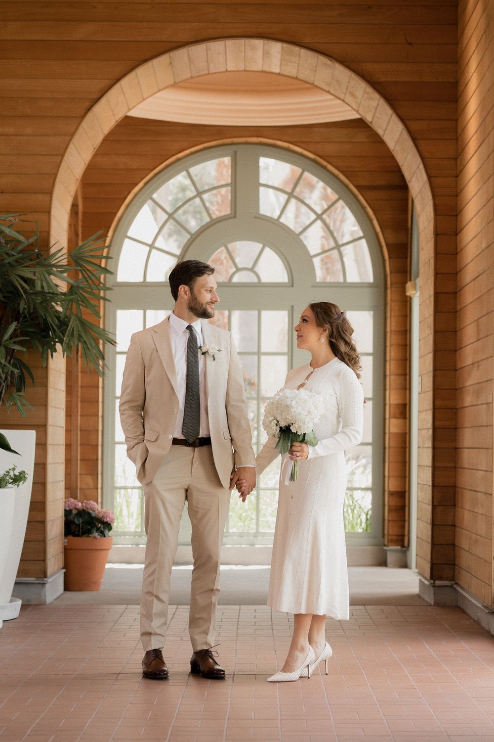 A couple in light colored formal attire stands in an elegant arched hallway with terracotta floors and wooden walls.