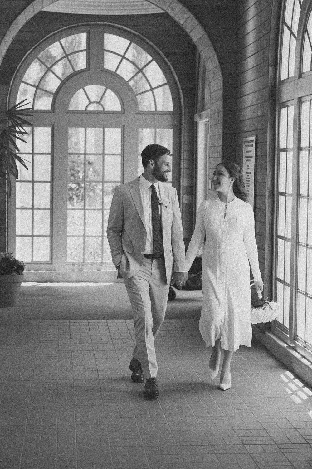 Two people walk through an elegant arched hallway with large windows casting dramatic shadows on the tiled floor.