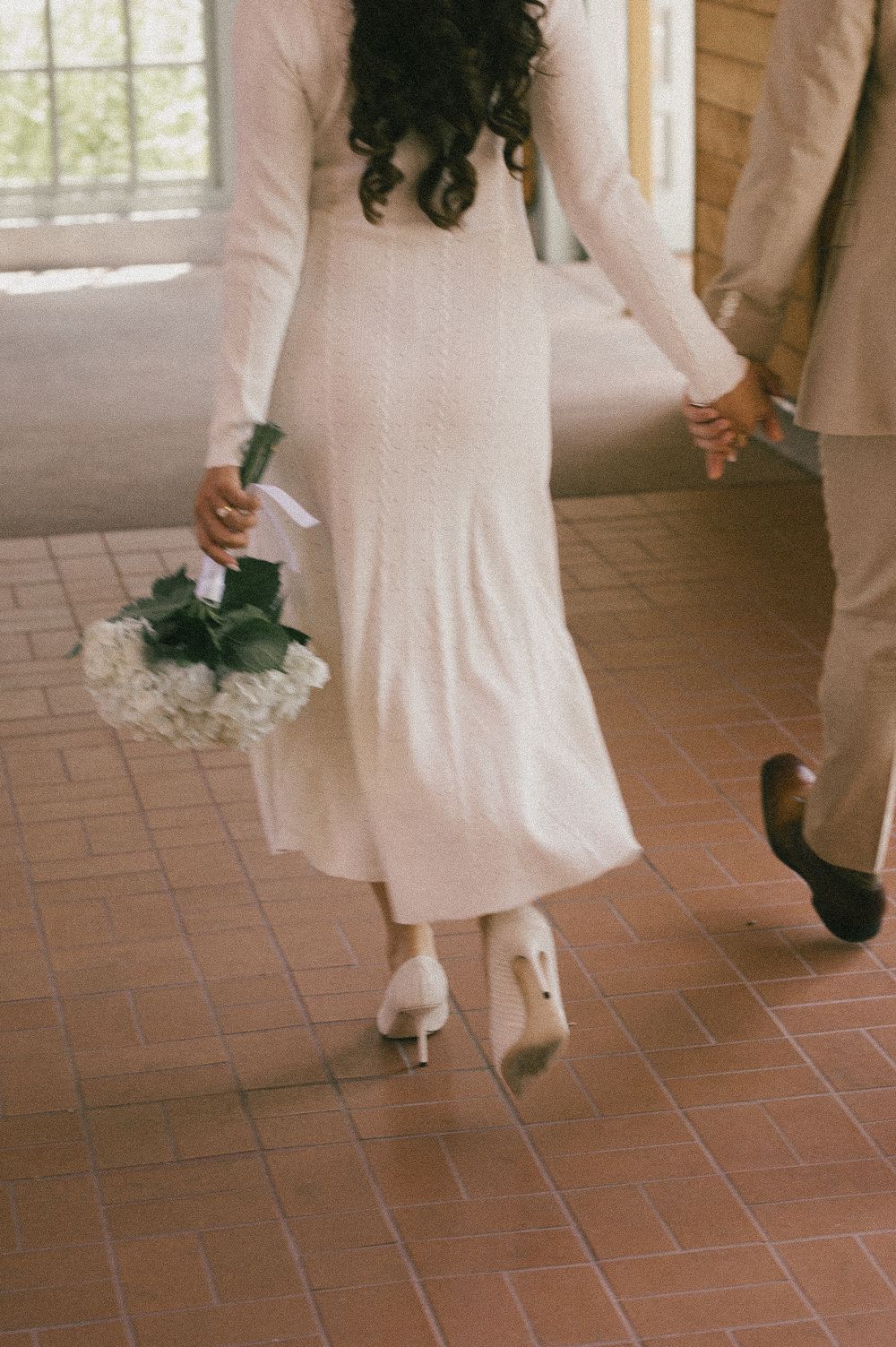 A person in a white dress walks across terracotta tiles, showing only the lower portion of their outfit and shoes.
