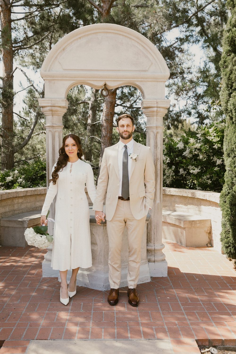 A couple in white attire poses in a Mediterranean-style courtyard with arched columns and brick flooring.