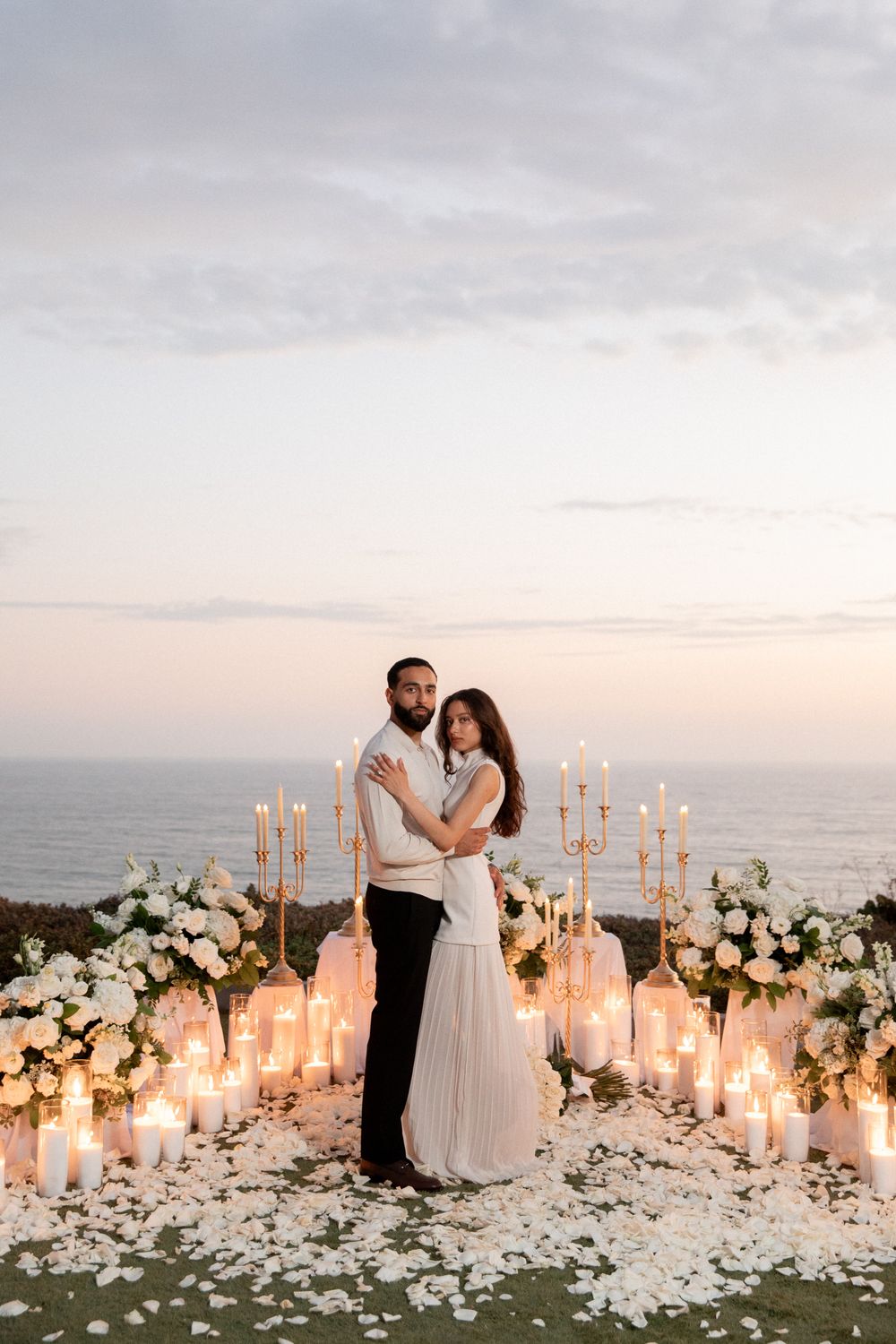 Romantic beach wedding ceremony setup with white flowers and glowing candles at sunset by the ocean.