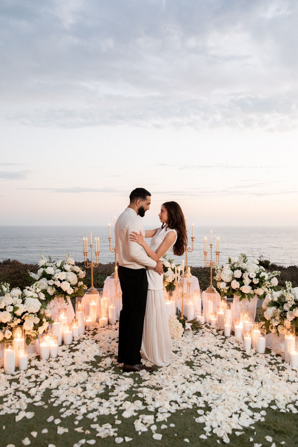 A romantic seaside wedding ceremony setup with white floral arrangements and glowing candles at sunset.