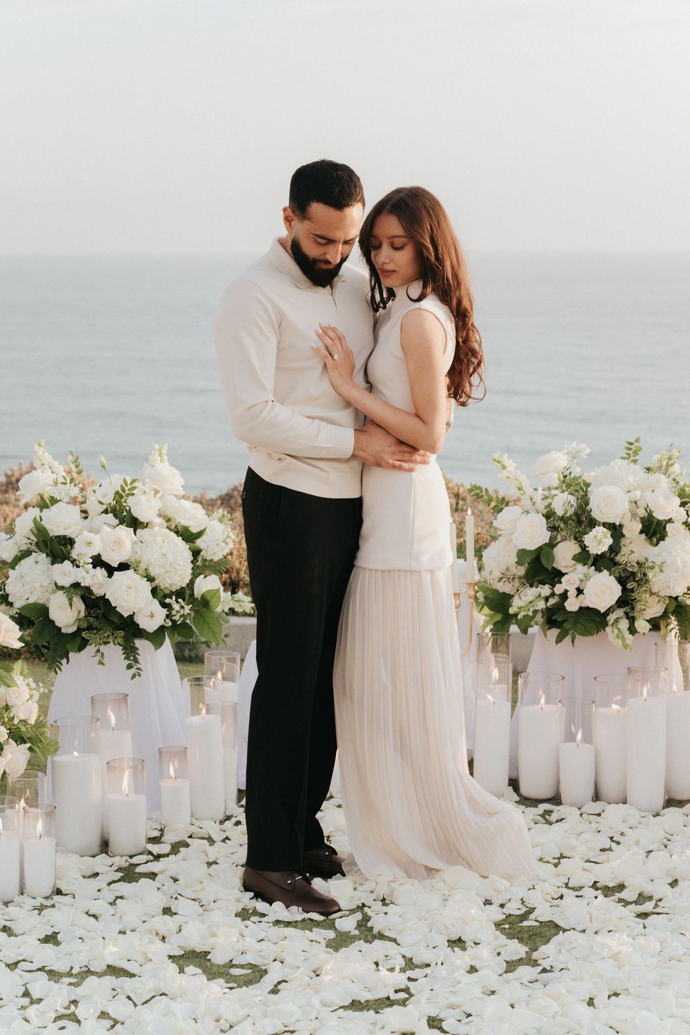Romantic beach wedding setup with white floral arrangements, candles and a couple embracing by the ocean.