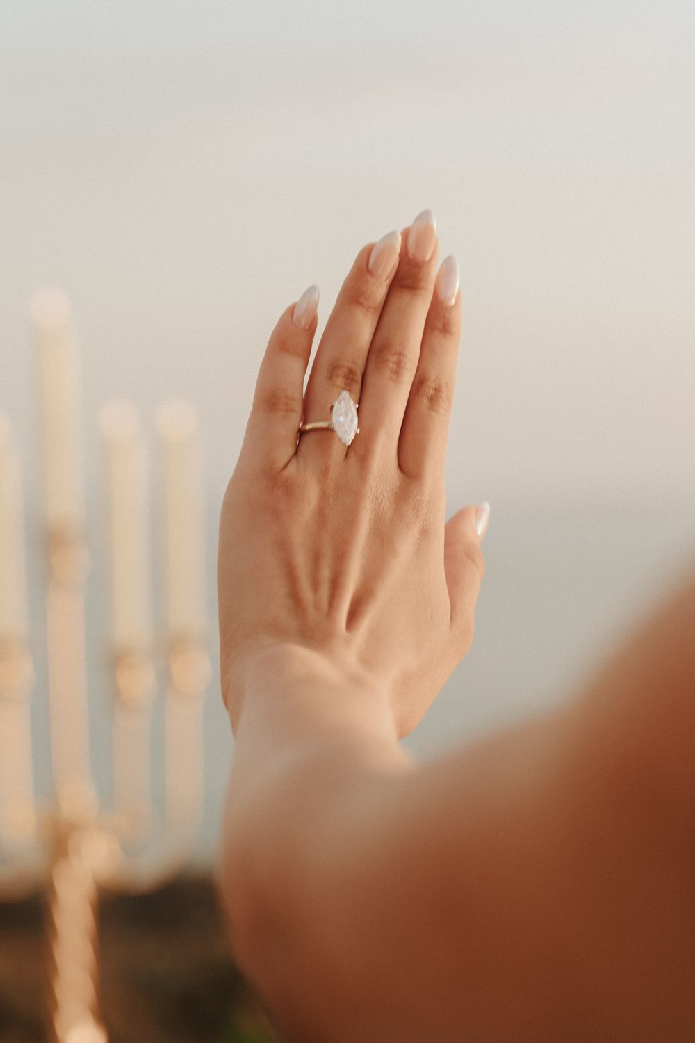 A hand with an engagement ring is raised against a blurry sunset background showing the sparkly diamond.