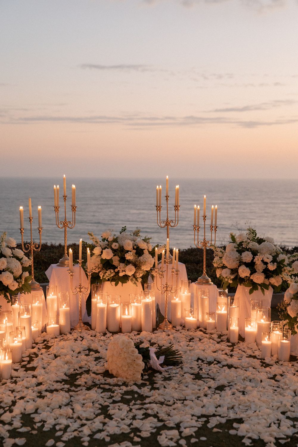 Romantic candlelit beach wedding setup with white flowers and golden candelabras at sunset overlooking the ocean.
