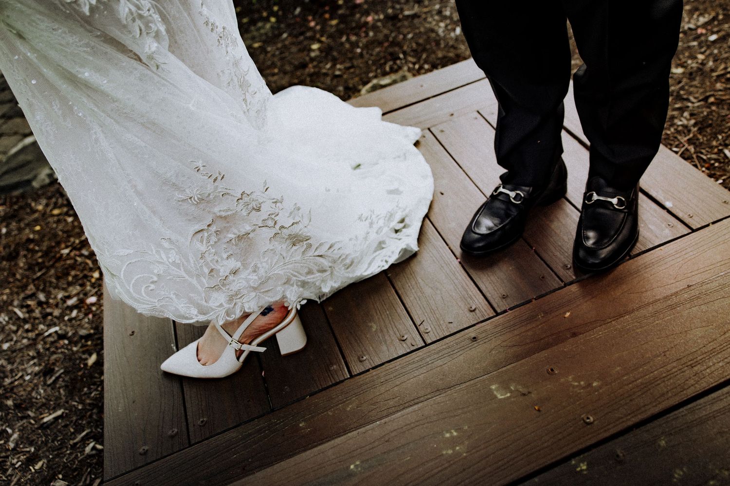 A close-up view of formal wedding shoes, with a white lace wedding dress and black dress shoes on wooden decking.