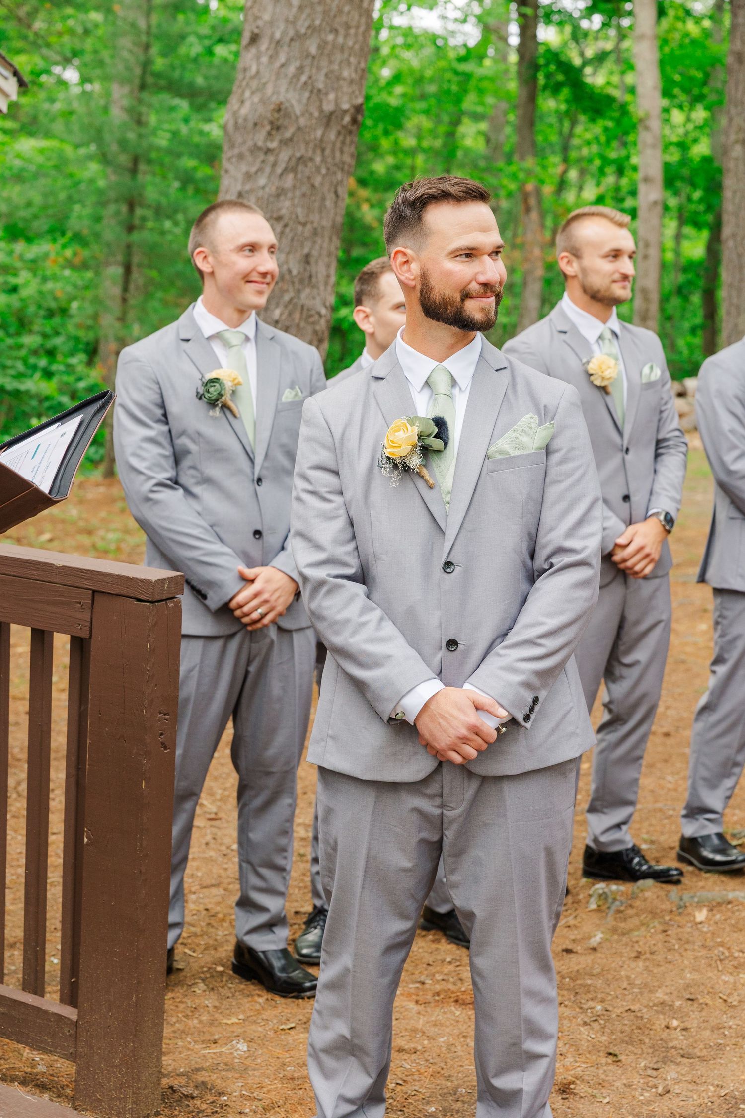 A couple walks down a wooded outdoor aisle during their summer wedding ceremony as guests look on.