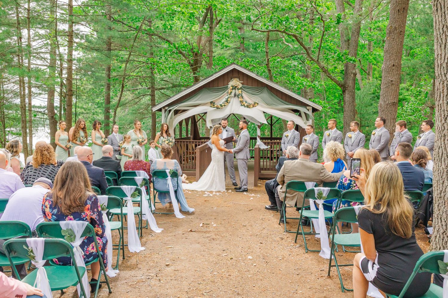 An outdoor woodland wedding ceremony takes place under a rustic wooden pavilion surrounded by green trees and seated guests.