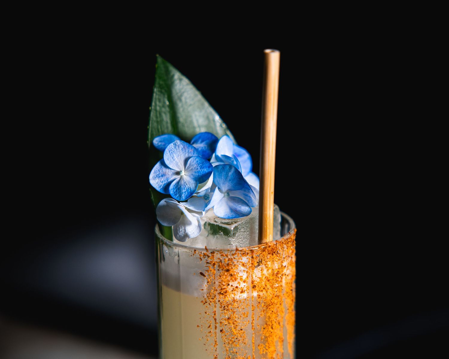 A tropical cocktail garnished with blue flowers and an orange-dusted rim against a black background.