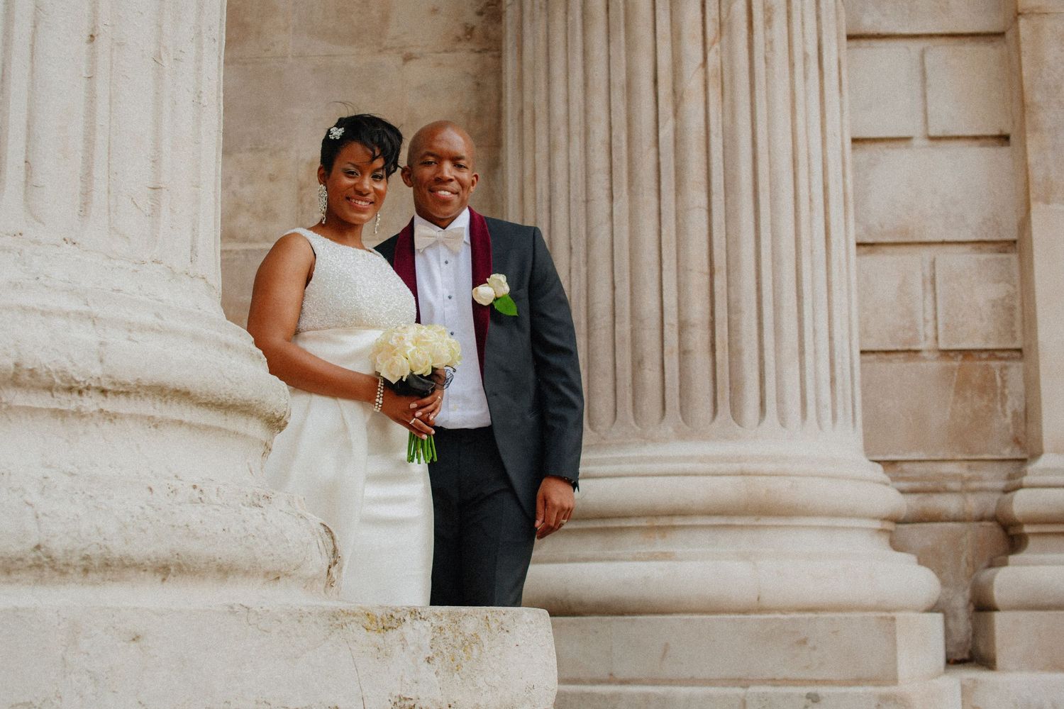 Bride and groom pose between imposing columns at St Paul's Cathedral. Bride in white strapless gown, groom in black suit