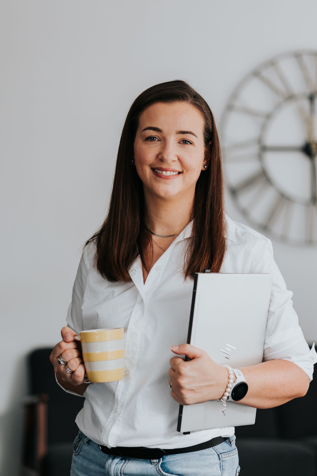 Professional in a white shirt holds a laptop and striped coffee mug while standing in front of a wall clock.