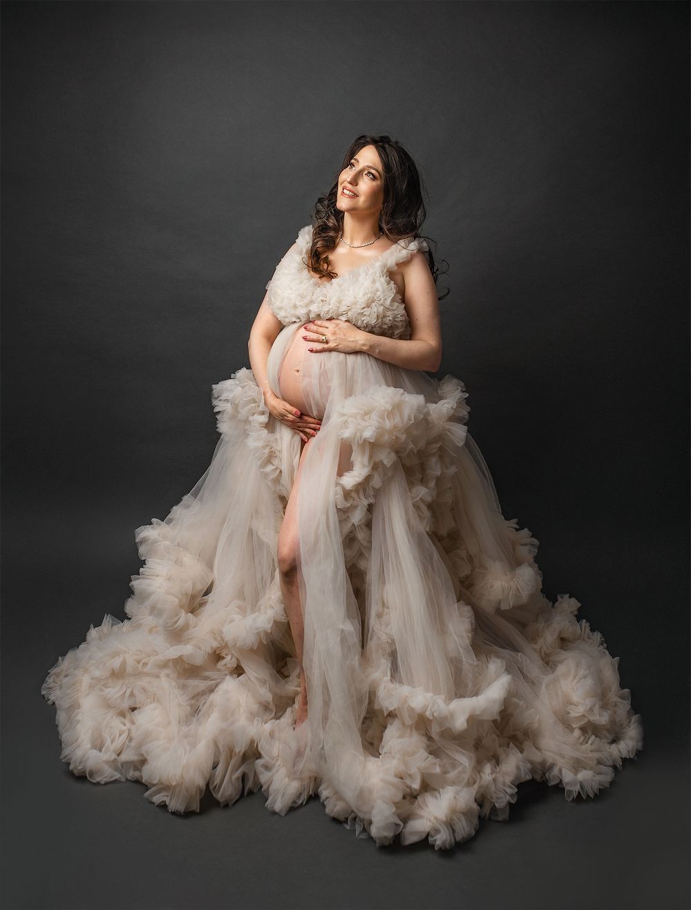 An expectant mother in a flowing ivory tulle gown with ruffled details poses against a dark studio backdrop.