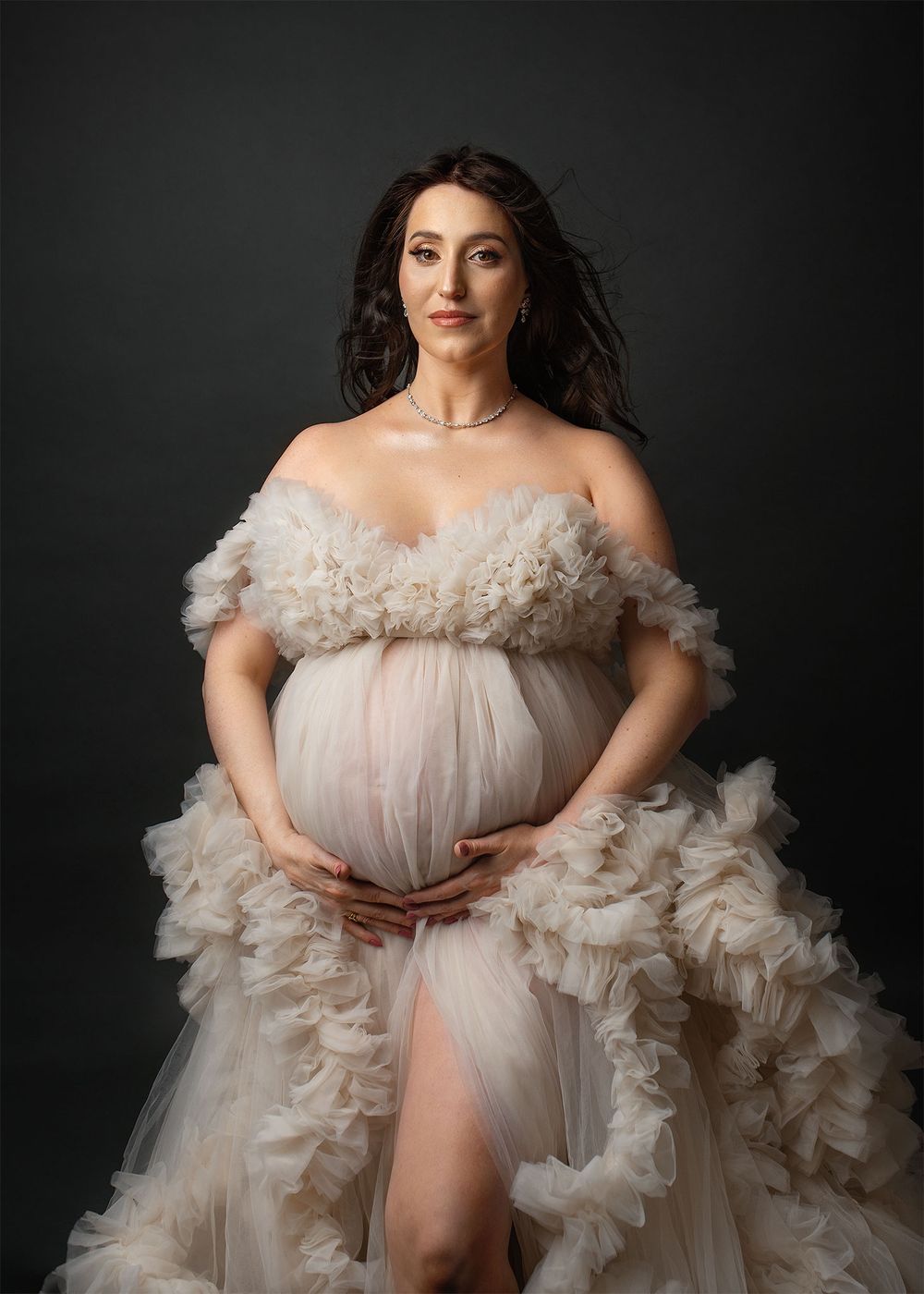 Ethereal maternity portrait with flowing cream-colored tulle gown against dark background in dramatic studio lighting.