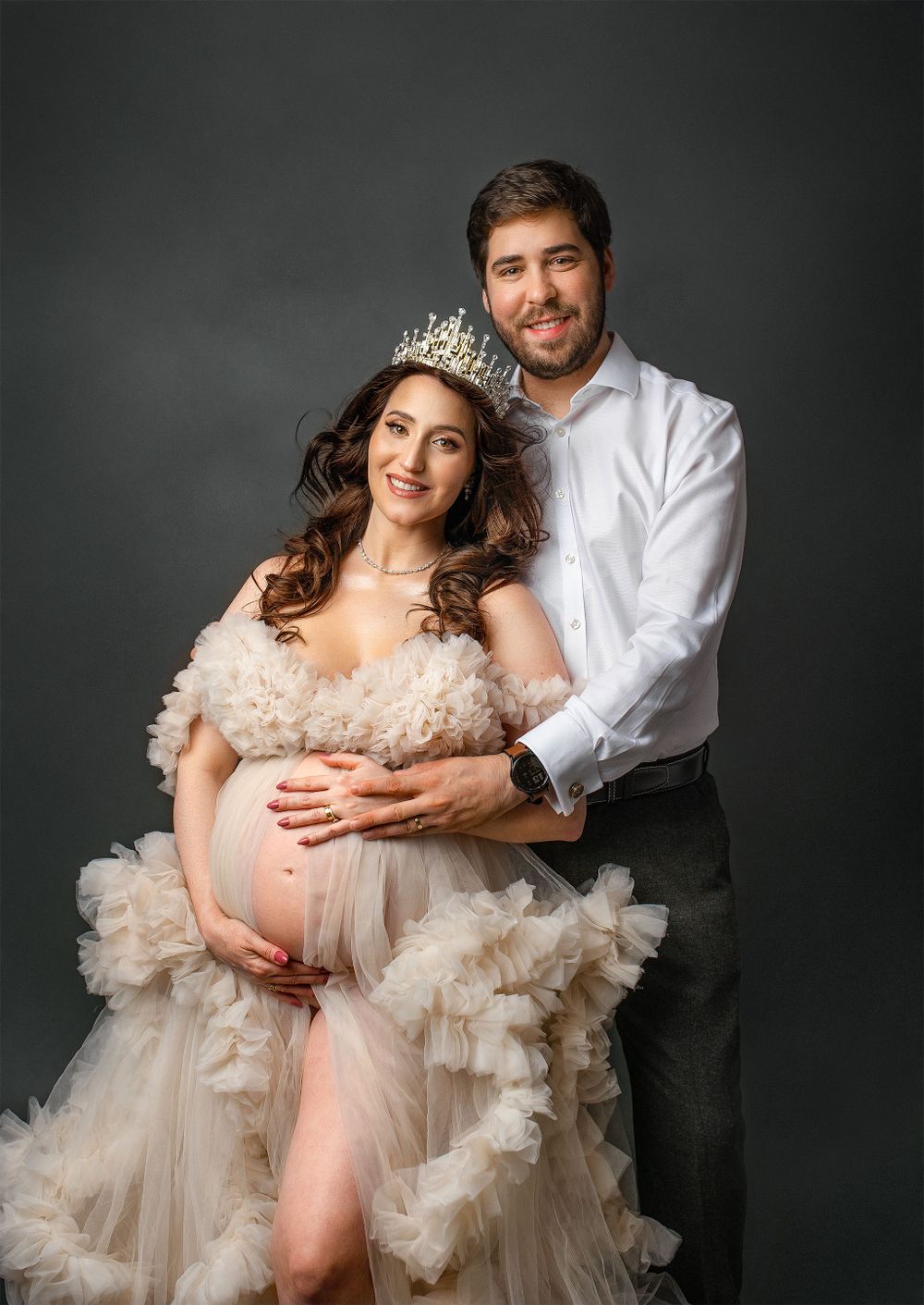 Maternity portrait with a couple in white together against dark background, wearing formal attire with tiara and tulle gown.