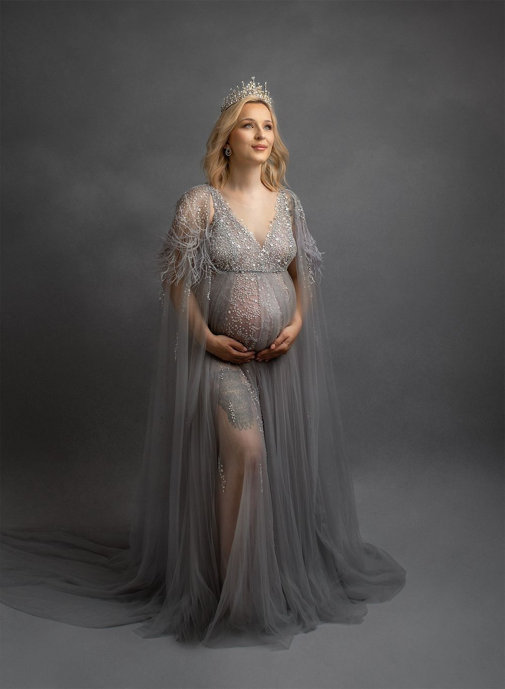 Ethereal maternity portrait in silver gray tulle gown and crown against dark backdrop creating dreamy atmosphere.