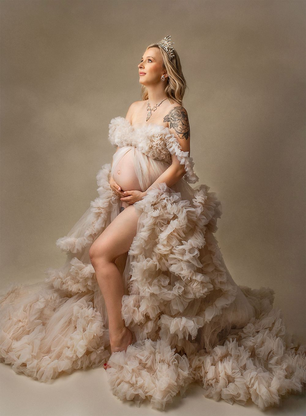 Elegant maternity photo featuring a flowing cream tulle gown and delicate crown in a ethereal studio setting.