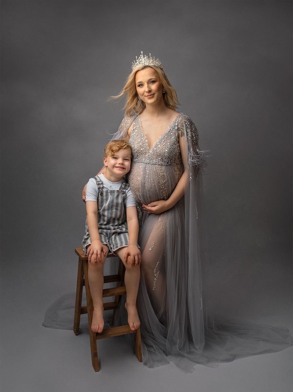 A maternity portrait with a pregnant figure in a flowing sheer gray gown and tiara with her son sitting on a wooden stool.