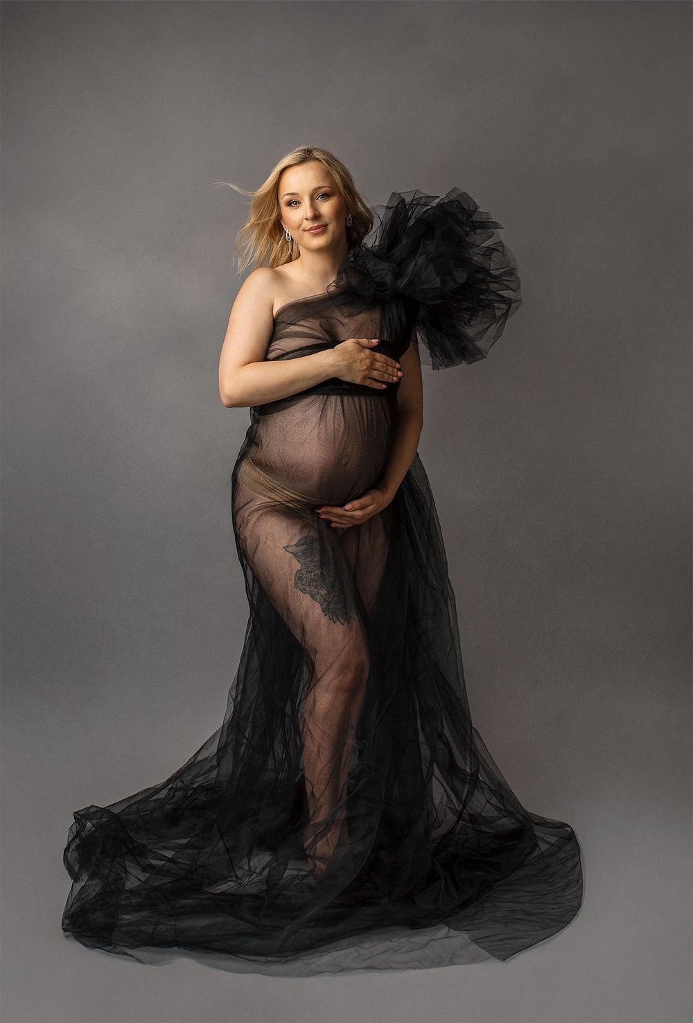 Pregnant figure poses in elegant sheer black tulle gown with dramatic flowing fabric against gray studio background.