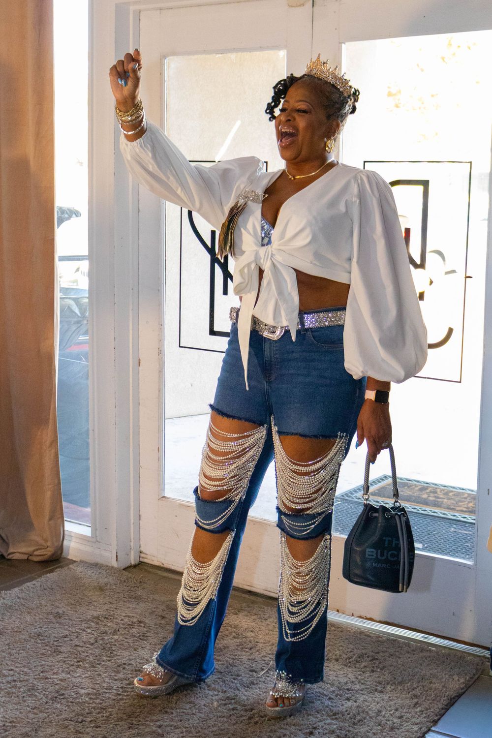 Woman in white crop top, ripped denim flares, and tiara poses confidently at a doorway.